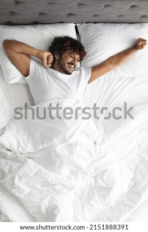 Happy curly hindu guy in white t-shirt stretching body while laying in comfortable bed, looking at copy space and smiling, enjoying new day, feeling fully rested, top view, vertical shot Royalty-Free Stock Photo #2151888391