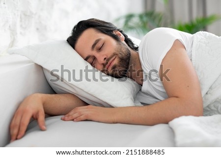 Healthy sleep concept. Closeup of handsome bearded young man sleeping in nice comfortable bed at home, enjoying good sleep, side view, copy space, white bedroom interior Royalty-Free Stock Photo #2151888485