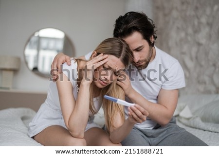 Upset young husband comforting his depressed young wife with negative pregnancy test on bed at home. Millennial couple cannot have baby, suffering from infertility indoors Royalty-Free Stock Photo #2151888721