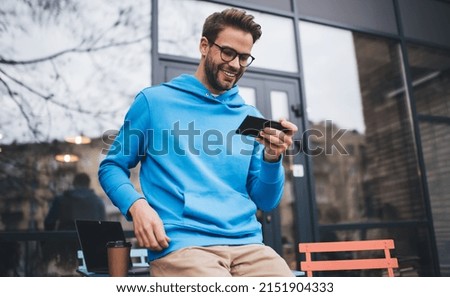 Millennial male blogger in classic spectacles connecting to 4g internet on digital cellular gadget for making online booking on website, cheerful hipster guy installing application on smartphone Royalty-Free Stock Photo #2151904333
