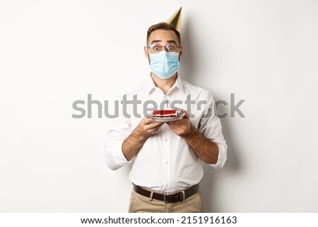 Covid-19, social distancing and celebration. Surprised birthday guy holding bday cake, wearing face mask from coronavirus, white background