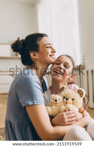 Smiling mother hugging cute little girl holding teddy bear toy, both laughing while cuddling, girl sitting on mom's knees, female tickling her child. Human relationships. Carefree childhood Royalty-Free Stock Photo #2151919549