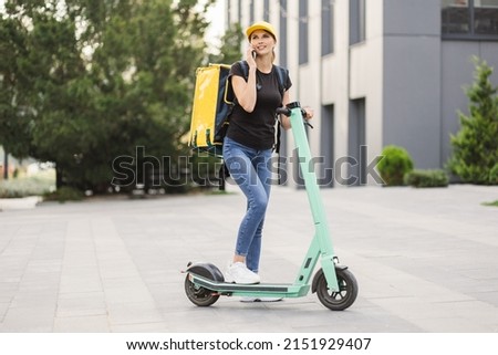 Delivery woman using smartphone while standing in the city with electric push scooter. Adult delivery woman in black t-shirt and jeans delivering online order to client.