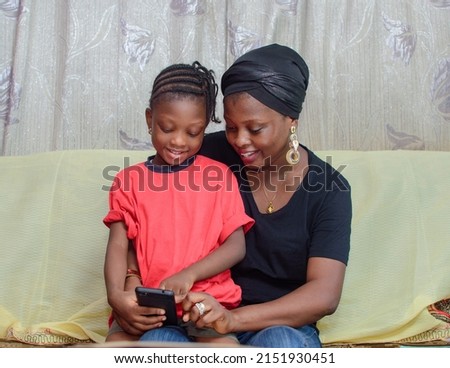 Lovely African Nigerian family consisting of a mother or guardian and her little girl child sitting on the mom's laps as they happily look at the smart phone in their hands