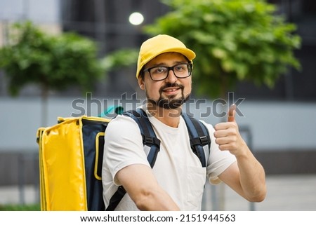 Delivering Food. Close up portrait of courier man with yellow backpack smiling to camera on the background of modern buildings, outside showing thumb up.