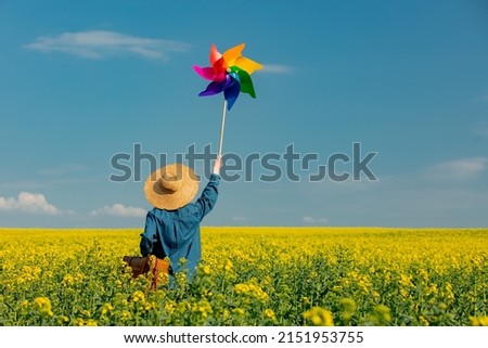 Woman in hat with suitcase and pinwheel in rapeseed field Royalty-Free Stock Photo #2151953755
