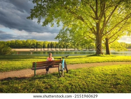 Woman sitting on bench and mountain bike, green trees and lake at sunset in spring. Colorful landscape with resting girl, bicycle, river, green grass, river in park. Summer. Sport and travel. Biking Royalty-Free Stock Photo #2151960629
