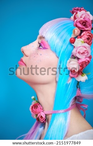 Beauty, makeup and hairstyle. Sideview portrait of a pretty teen girl with bright pink make-up posing in colored violet-blue wig and flower wreath on the head. Studio portrait on a blue background.