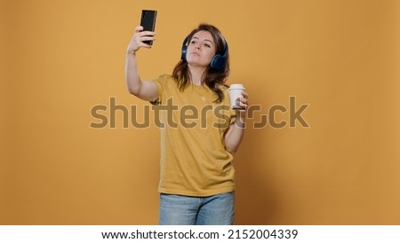 Portrait of relaxing woman taking a selfie while holding a cup of coffee to go while listening to music on wireless headphones in studio. Person using smartphone to take picture using front camera.