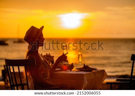 Romantic dinner near sea on sunset. Woman sitting alone, waiting husband on table set for a romantic meal on beach sky and ocean on background. Dinner for a couple in love in luxury outdoor restaurant Royalty-Free Stock Photo #2152006055