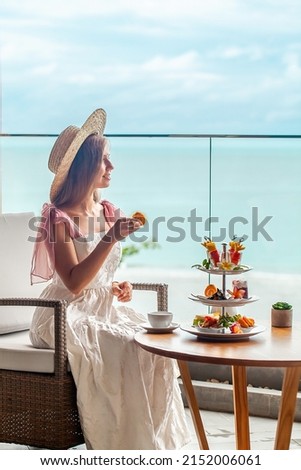 Luxury lifestyle. Elegant young lady in white dress and straw hat eating cake and enjoy sweet food from cake stand in cafe with sea view. Romantic afternoon tea in restaurant Royalty-Free Stock Photo #2152006061