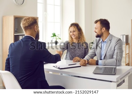 Happy married couple shaking hands with realtor after signing papers at his office. Clients exchanging handshake with real estate agent, smiling and thanking him for professional consultation and help