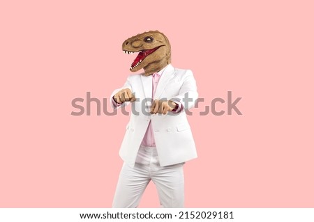 Weird guy in funny disguise dancing against pastel pink studio background. Cheerful eccentric man in white suit and silly ugly wacky masquerade dinosaur mask having fun at crazy party Royalty-Free Stock Photo #2152029181