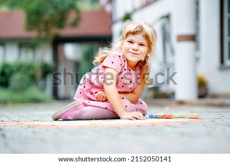 Little preschool girl painting rainbow with colorful chalks on ground on backyard. Positive happy toddler child drawing and creating pictures. Creative outdoors activity in summer.