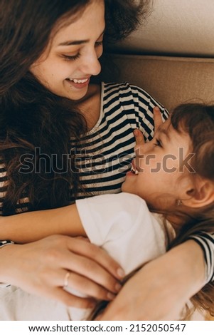 Vertical close-up of cute little girl lying in hands of her young beautiful charming mother, telling her stories, before going to bed, mom smiling at her daughter. Strong bonds. Human relationships