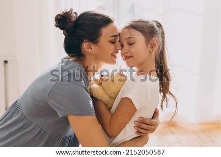 Picture of beautiful caring young mother hugging her cute little daughter tight, holding teddy bear with eyes closed, embracing her child gently, showing her love, protection and admiration