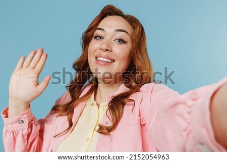 Close up friendly young redhead chubby overweight woman 30s in pink shirt doing selfie shot on mobile phone waving hand isolated on pastel blue background studio portrait. People lifestyle concept.