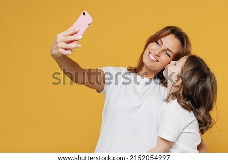 Happy woman in basic white t-shirt have fun doing selfie cute child baby girl 5-6 years old. Mom little kid daughter isolated on yellow orange color background studio. Mother's Day love family concept