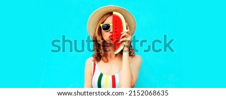 Summer portrait of young woman covering half of her face with slice of watermelon blowing her lips with red lipstick wearing straw hat on blue background, blank copy space for advertising text