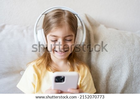 Cute child with headphones and mobile phone on sofa indoors.Smiling small child playing online game on smartphone at home, resting on sofa.
