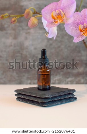 Mock-up of glass bottle of essential oil or serum for cosmetics with orchid flowers on gray background. Cosmetic bottle. Skin care and healthy facial and body care. Vertical photography