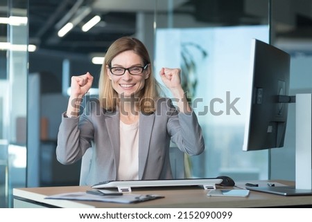 Beautiful and successful business woman with glasses, celebrates victory, looks at the camera and rejoices in a successfully completed project, keeps her hands up from happiness and satisfaction Royalty-Free Stock Photo #2152090099