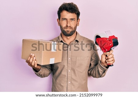 Handsome man with beard holding packing tape holding cardboard skeptic and nervous, frowning upset because of problem. negative person. 