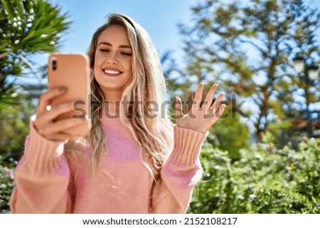Young blonde woman doing video call waving to the camera at the park