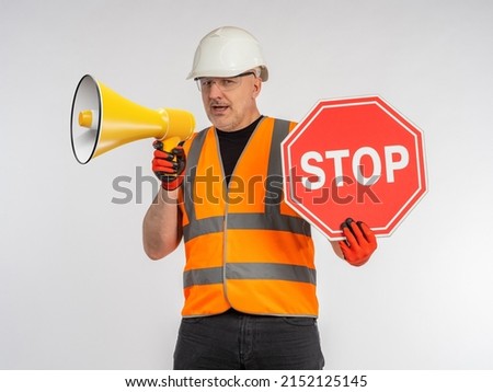 Man in road worker's uniform. Builder with loudspeaker. Road worker shows stop sign. Portrait of road repairman with loudspeaker. Builder shouts into megaphone. Man builder on light background