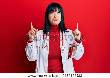 Young hispanic woman wearing doctor uniform and stethoscope pointing up looking sad and upset, indicating direction with fingers, unhappy and depressed. 