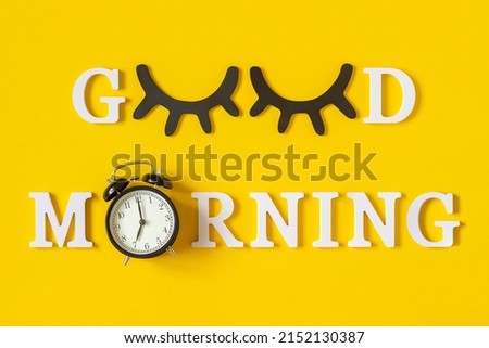 Creative good morning concept. White letters, black eyeleshes and alarm clock on yellow background. Top view, Flat lay. Greeting card.