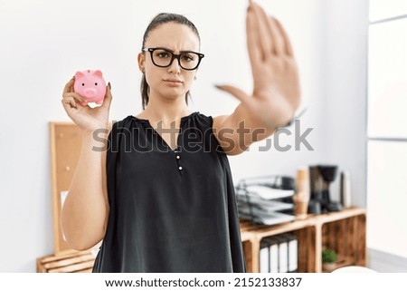 Young brunette woman holding piggy bank at the office with open hand doing stop sign with serious and confident expression, defense gesture 