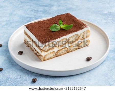 Tiramisu cake decorated with cocoa powder and fresh green mint leaf. Close up food. Blue background. Traditional italian dessert. Piece of cake on white ceramic plate. Royalty-Free Stock Photo #2152152547