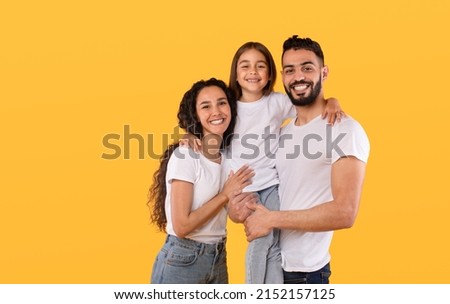 Arabic Parents And Daughter Embracing Together Posing Standing In Studio On Yellow Background, Wearing White T-Shirts. Shot Of Loving Middle Eastern Family Concept