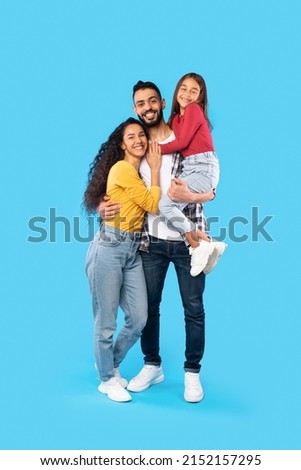 Arabic Man Hugging Wife And Holding Little Daughter In Arms Standing On Blue Background. Happy Middle Eastern Family Posing In Studio Smiling To Camera. Vertical Shot