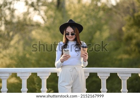 Beautiful young brunette girl in a hat with glasses and with a phone in her hands. Drinking coffee in the summer.