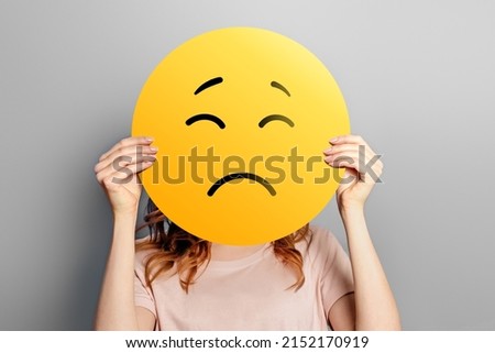 Sad emoji. Girl holds a yellow emoticon with sad face isolated on a gray background. unhappy emoji