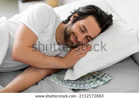 Smiling bearded young guy sleeping with bunch of cash under his pillow, keeping his money savings by him at home, closeup photo. Finance management concept