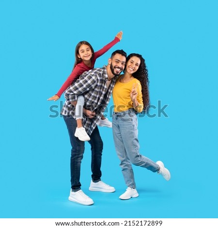 Cheerful Middle Eastern Family Having Fun Posing Together, Father Carrying His Daughter Piggyback Standing Over Blue Studio Background. Square Shot, Full Length Royalty-Free Stock Photo #2152172899
