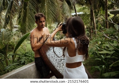 Beautiful girl photographer takes pictures of a surfer guy in a palm forest