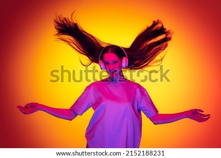 Music. Emotional caucasian girl wearingt-shirt dancing, listening to musci isolated on yellow-red background in neon. Concept of beauty, art, fashion, youth, sales and ads. Royalty-Free Stock Photo #2152188231