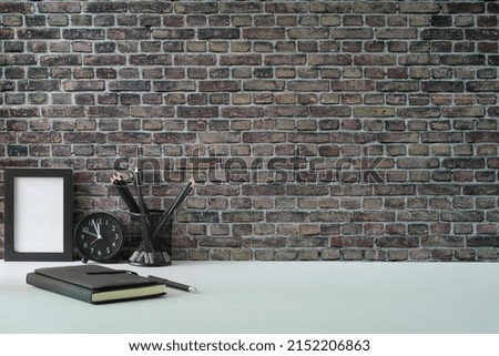 Stylish workplace. Empty picture frame, pencil holder and notebook on white table against brick wall with copy space.