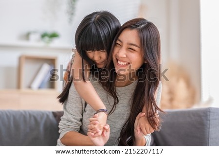 Fun mom with cute baby girl. Mother and daughter playing together in the living room. Asian girl embracing mom from stand behind while mom sitting on couch. Happy family, motherhood, childhood concept