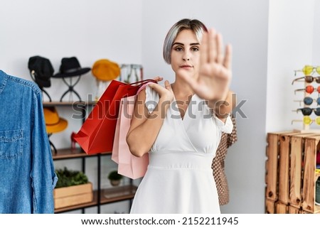 Young beautiful woman holding shopping bags at retail shop with open hand doing stop sign with serious and confident expression, defense gesture 