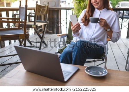 Closeup image of a young woman holding and using mobile phone while drinking coffee with laptop computer on the table