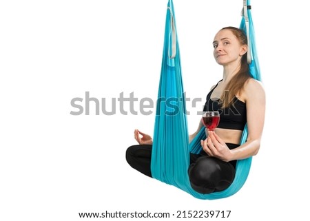 Young girl hangs in hammock sitting in lotus position and holding glass of red wine. Sports, meditation and alcohol concept