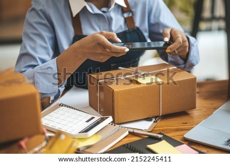 A woman using a smartphone to take pictures in front of parcel boxes, parcel boxes for packing goods, delivering goods through private courier companies. Online selling and online shopping concepts.