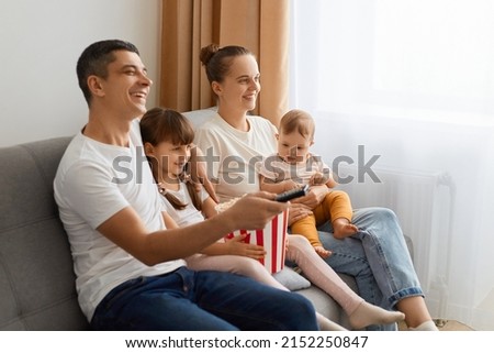 Side view portrait of family sitting on sofa with remote control and watching tv, enjoying interesting film or cartoon, being extremely happy being together at home.