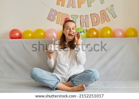 Image of delighted satisfied attractive young woman wearing white shirt celebrating birthday, sitting alone on sofa with cake, blowing party horn, looks extremely happy.