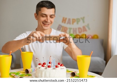 Indoor shot of smiling brunette young adult man wearing white t shirt sitting at table with his smart phone and taking picture, photographing his birthday cake.